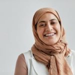 A cheerful Muslim woman wearing a hijab smiles brightly against a white backdrop.